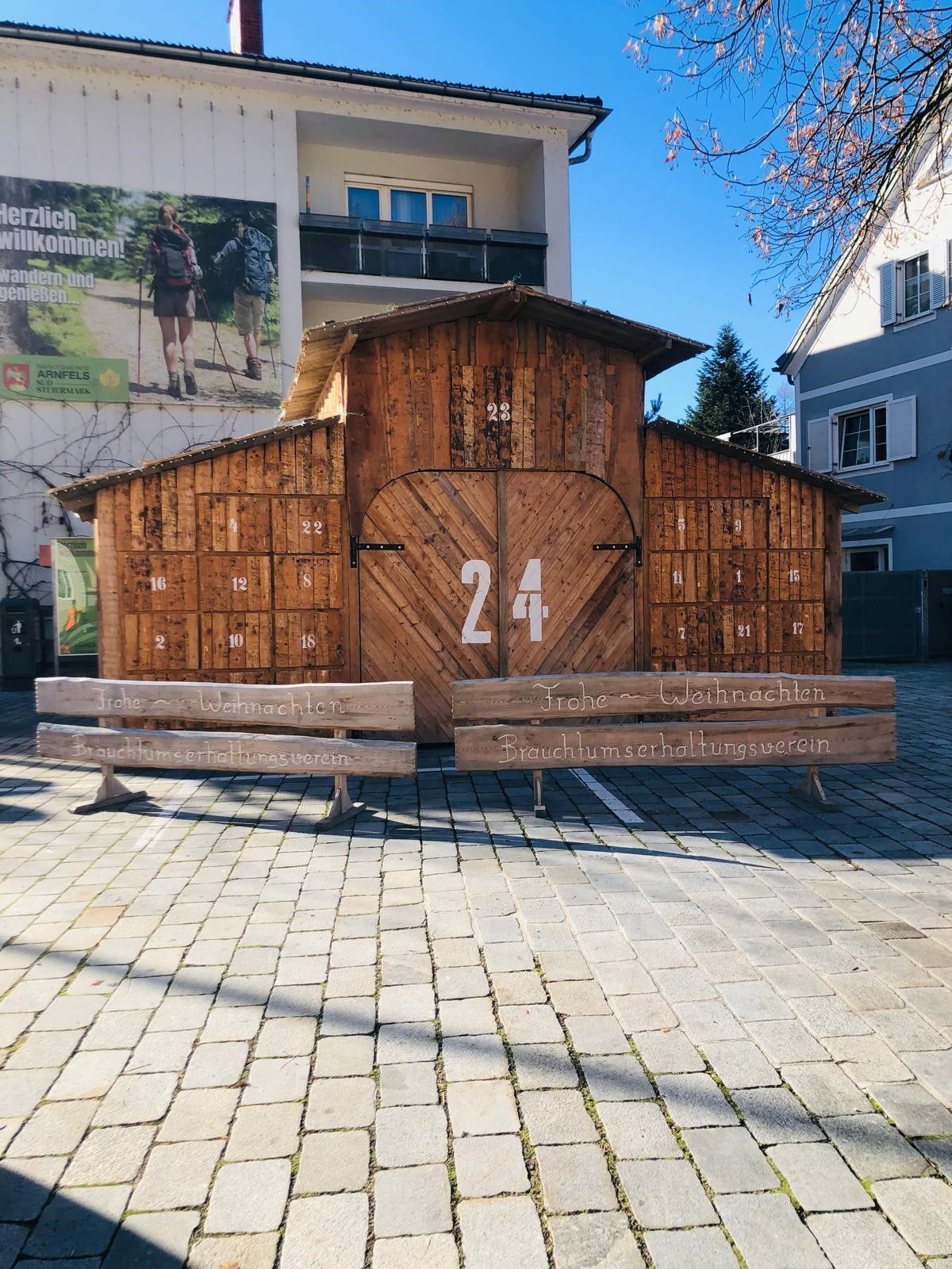 Eine Holzstruktur mit einer Tür, die mit 24 nummeriert ist, und Bänken mit den Aufschriften 'Frohe Weihnachten' und 'Brachunserhaltunsverein' auf einem Kopfsteinpflaster. Dahinter steht ein Gebäude mit einem Schild mit Wanderern.