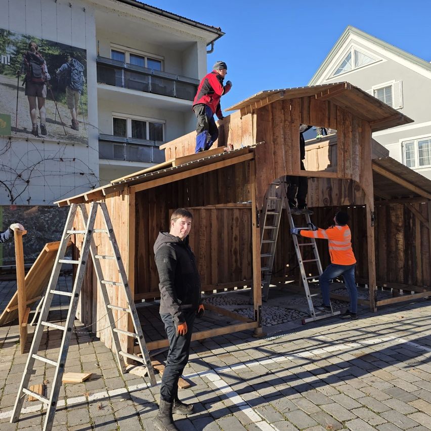 Drei Männer bauen eine Holzhütte auf einer Kopfsteinpflasterstraße. Einer steht auf dem Dach, ein anderer auf einer Leiter, und der dritte sieht zu. In der Nähe lehnt eine Leiter an der Hütte.
