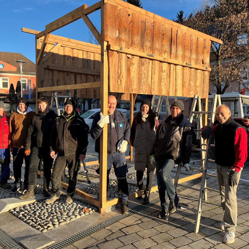 Eine Gruppe von Menschen steht um eine Holzstruktur auf einem Kiesweg, einige tragen Winterkleidung und Handschuhe. Ein Mann lehnt sich an die Struktur, ein anderer hält eine Leiter. Hinter ihnen sind Gebäude und ein geparktes Auto.