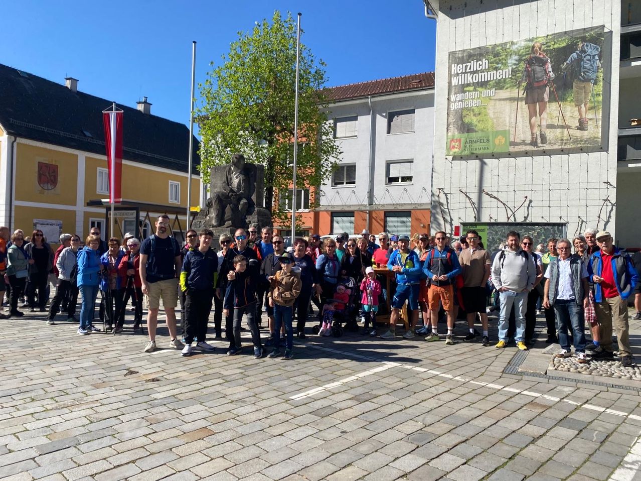 A group of people stand in front of a building with a statue and a sign that reads Herzlich willkommen! Wandern und genieen. They are all dressed for outdoor activities.