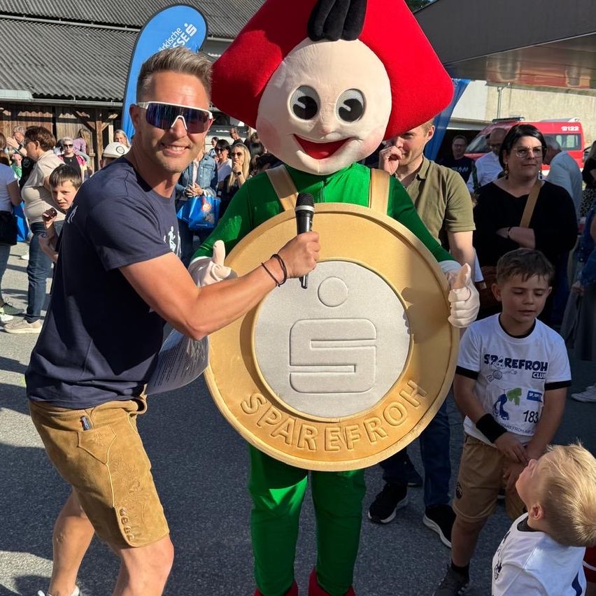A man holds a mascot's hand and a microphone. The mascot wears green and red with a gold coin featuring the number 5. They stand among a crowd.
