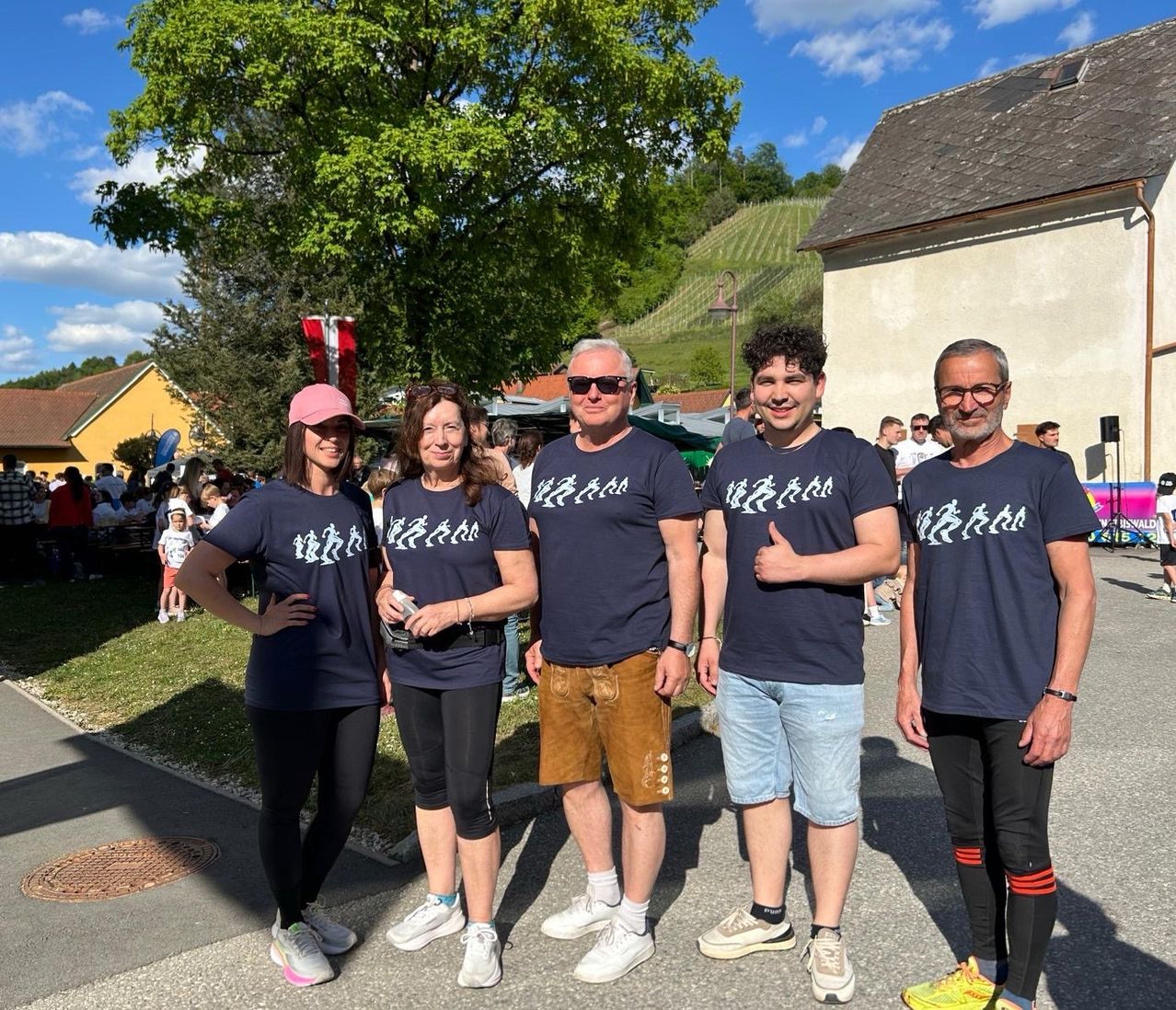 Five people wearing matching T-shirts with running figures are posing for a photo. They stand on a street with a building, tree, and hill in the background.