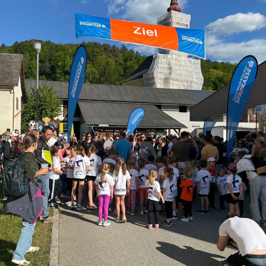 A crowd of people, including children in matching shirts, gather in front of a building with a tower, under a banner reading 'Ziel'.
