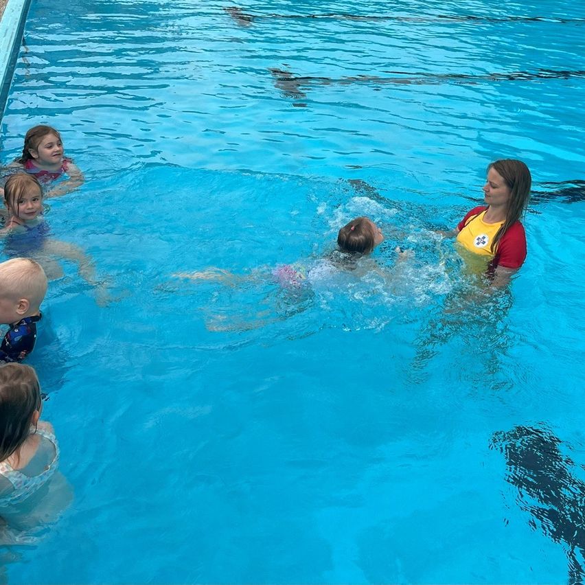 Eine Frau unterrichtet kleine Kinder im Schwimmen in einem Pool. Die Kinder tragen Badeanzüge und die Frau trägt eine gelbe Rettungsweste.