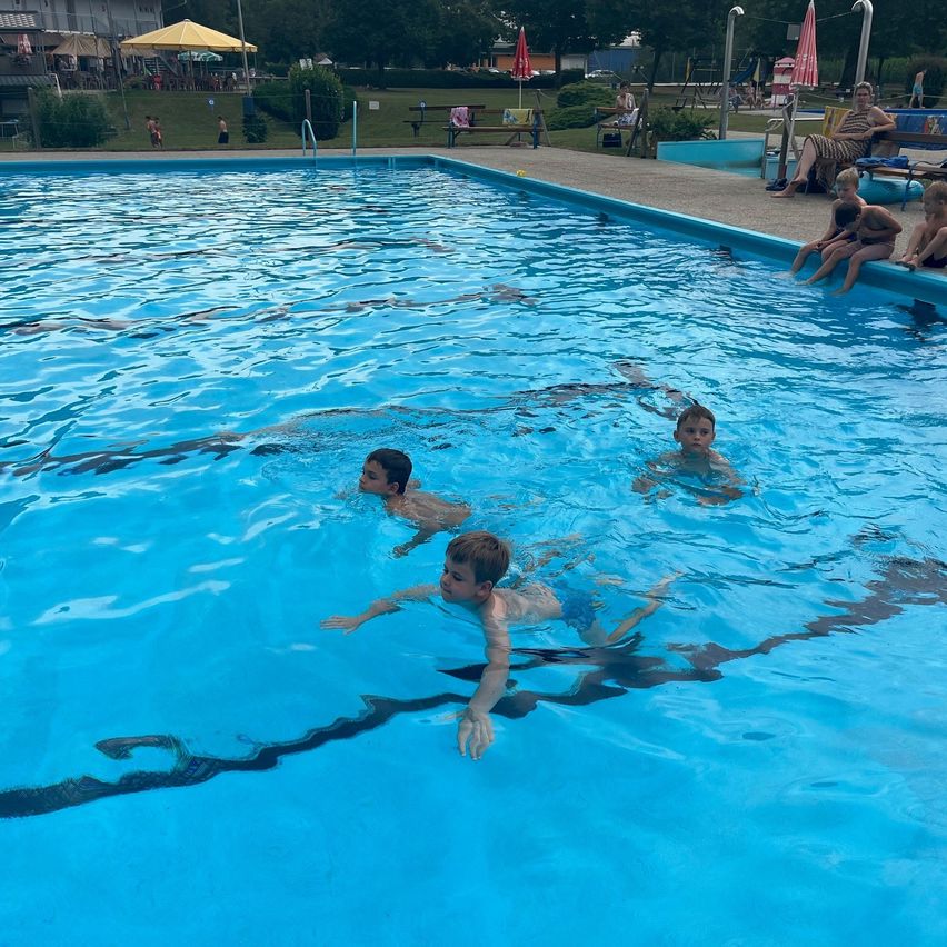 Drei junge Jungen schwimmen in einem großen Freibad mit klarem blauem Wasser. Eine Frau und zwei Kinder sitzen auf der Poolbank.