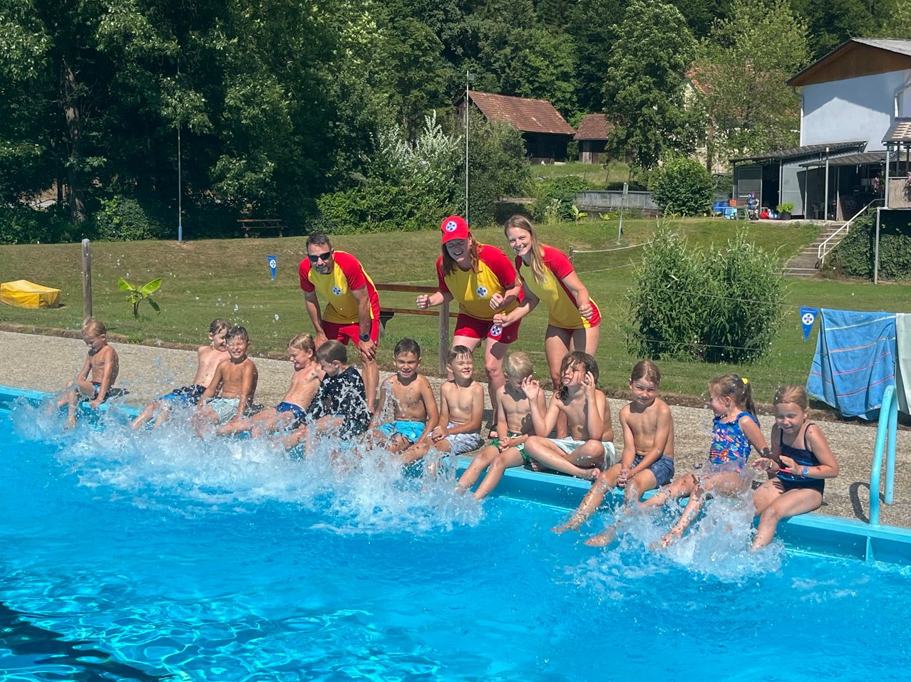 Eine Gruppe von Kindern und Erwachsenen in roten und gelben Uniformen sitzt in einem Pool, mit zwei Erwachsenen vor ihnen. Die Kinder spritzen Wasser und die Erwachsenen lächeln. Dahinter befindet sich ein Rasenbereich mit einem Holzzaun, einem Haus und Bäumen.