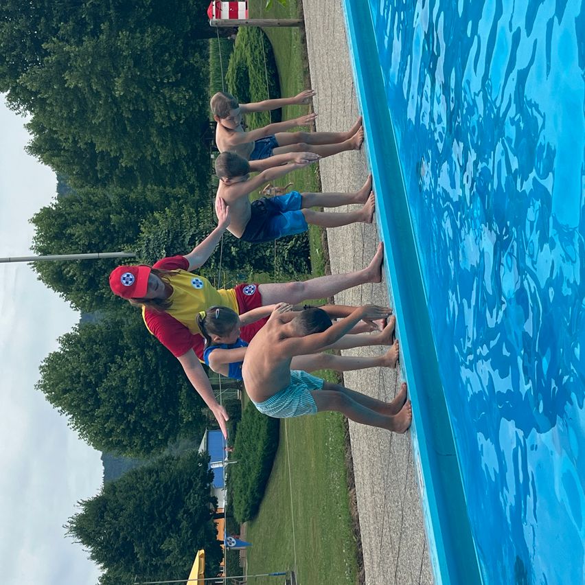 Eine Gruppe von Kindern und einem Erwachsenen stehen am Rand eines Schwimmbeckens und bereiten sich darauf vor, hineinzuspringen.
