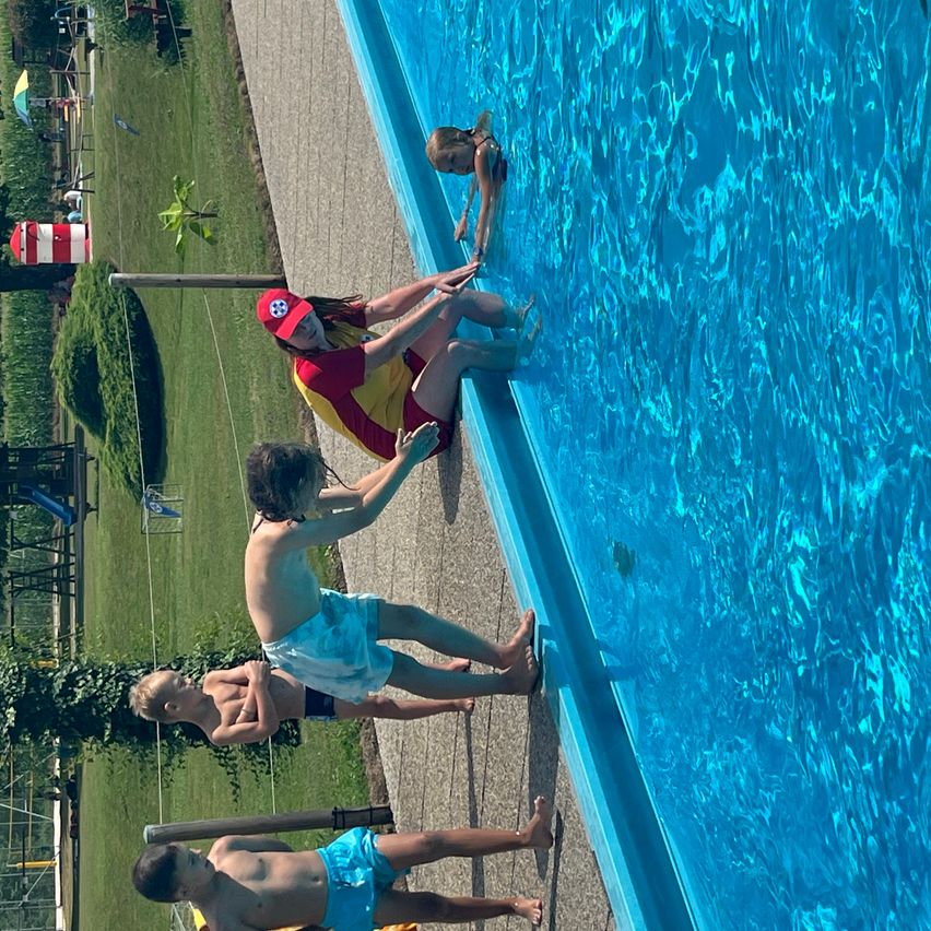 Eine Gruppe von Kindern und Erwachsenen versammelt sich um einen Pool, wobei eine Person im Wasser und andere am Rand stehen.
