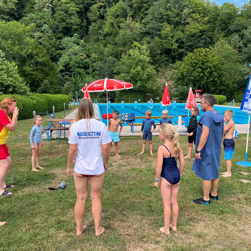 Eine Gruppe von Kindern und Erwachsenen versammelt sich in der Nähe eines Schwimmbeckens, wobei einige Badeanzüge tragen und andere Freizeitkleidung. Ein Erwachsener in einem weißen Shirt mit dem Wort 'Wasserrettung' steht vor ihnen. Der Pool ist von Bäumen umgeben und hat einen roten und weißen Schirm in der Nähe.