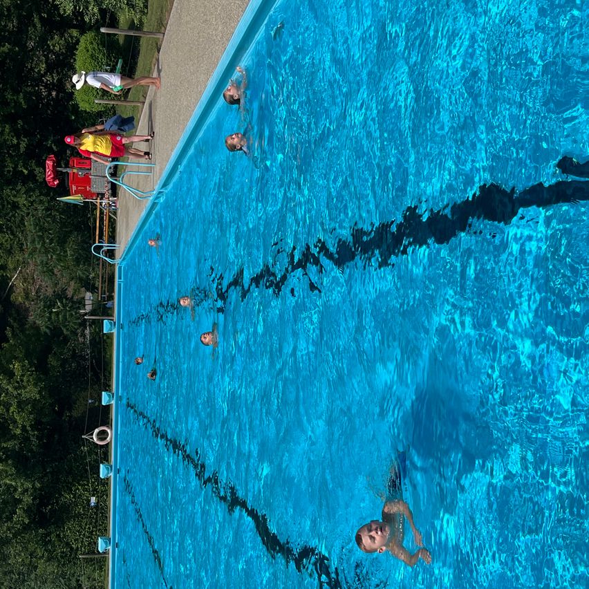 Eine Gruppe von Schwimmern in einem großen Freiluftbecken, wobei mehrere Personen in Bahnen schwimmen und andere auf der Poolterrasse stehen.