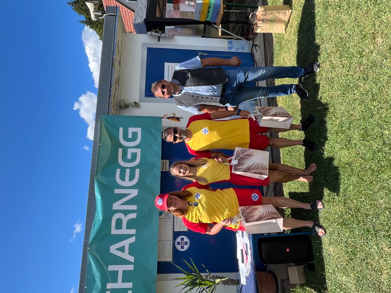 Vier Personen posieren für ein Foto vor einem Gebäude mit einem Banner, das 'Charnegg' sagt. Drei tragen passende gelbe und rote Outfits, während der vierte Mann in einem blauen Hemd und Jeans ist.