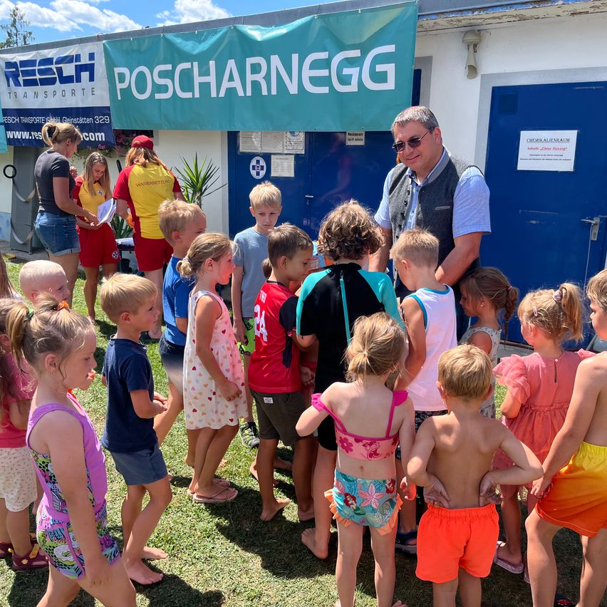 Eine Gruppe von Kindern und Erwachsenen versammelt sich vor einem Gebäude mit einem Banner, auf dem Poscharnegg steht. Ein Mann spricht mit den Kindern.