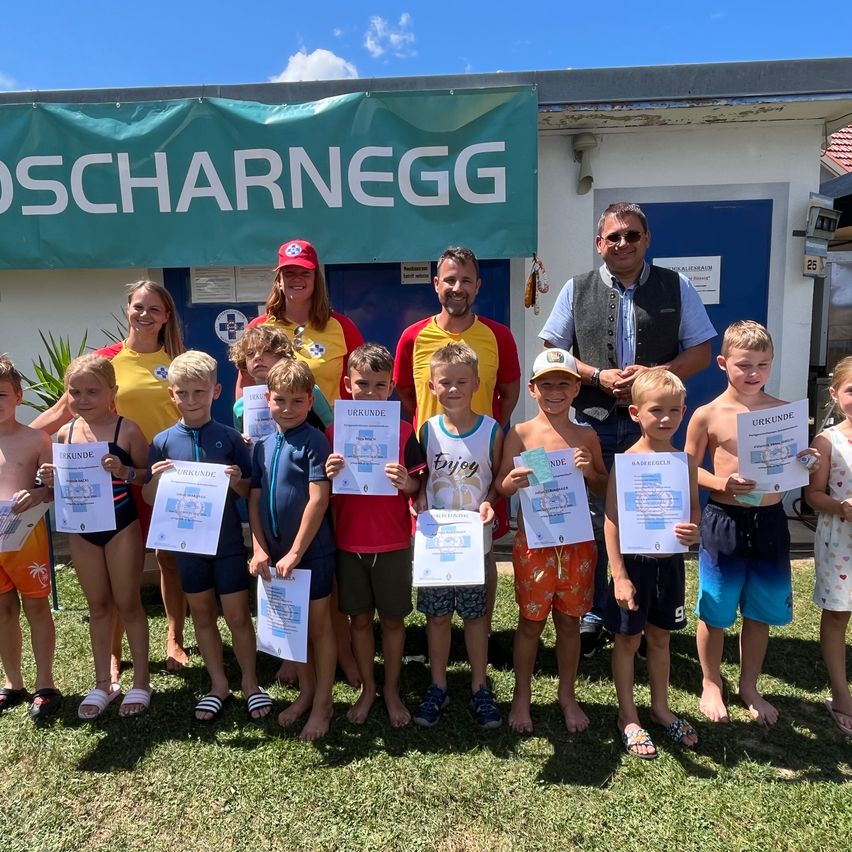 Eine Gruppe von Kindern und Erwachsenen steht in einer Reihe, hält Urkunden in der Hand und lächelt für ein Foto vor einem Gebäude mit einem Banner, das 'Oscharnegg' liest.