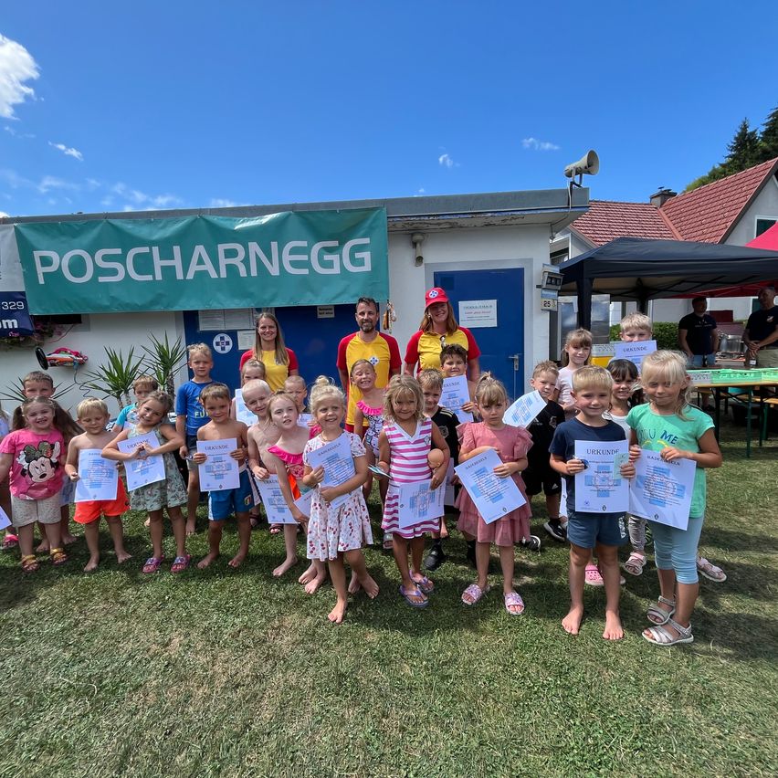 Eine Gruppe von Kindern und Erwachsenen steht auf einem Rasenfeld, hält Urkunden in der Hand, mit einem Gebäude und einem Banner im Hintergrund.