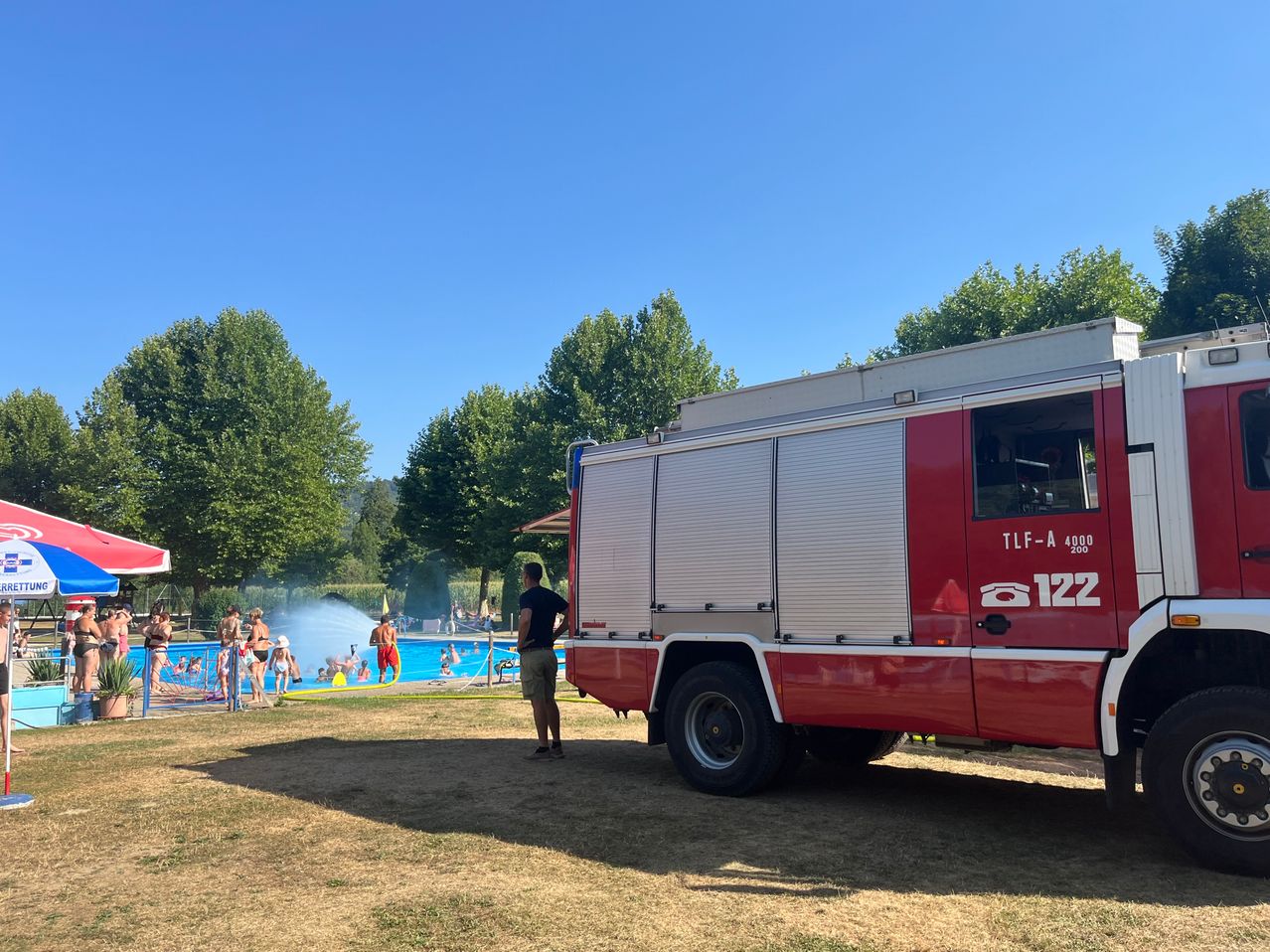 Ein Feuerwehrauto steht in der Nähe eines Schwimmbeckens, in dem Menschen schwimmen und am Rand stehen.