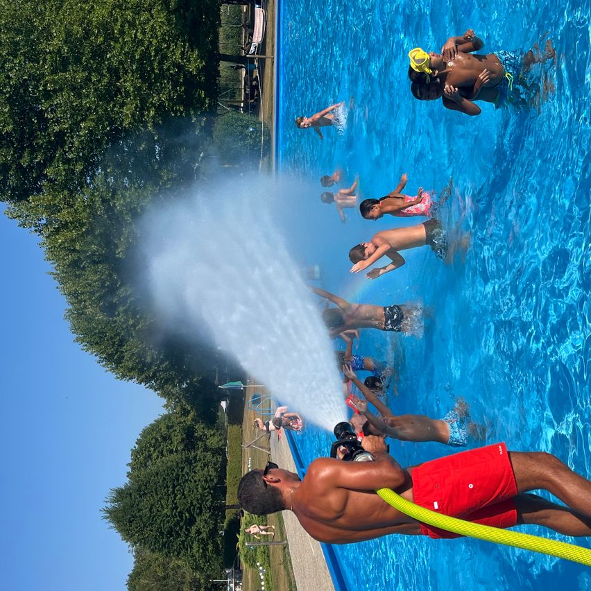 Eine Gruppe von Kindern und Erwachsenen schwimmt in einem Pool mit einer Wasserfontäne, die Wasser sprüht. Bäume und ein blauer Himmel sind im Hintergrund zu sehen.