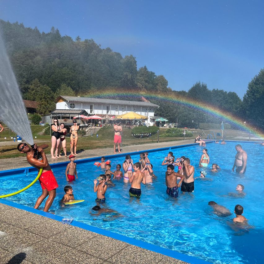 Eine Gruppe von Menschen genießt einen sonnigen Tag an einem Freibad, mit einem Regenbogen am Himmel und einem Gebäude im Hintergrund.