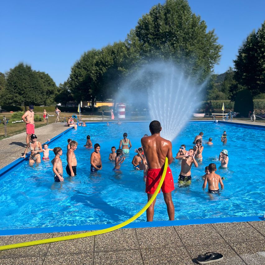 Eine Gruppe von Kindern und Erwachsenen schwimmt in einem großen Außenpool, während ein Mann einen Wasserschlauch hält und Wasser sprüht.