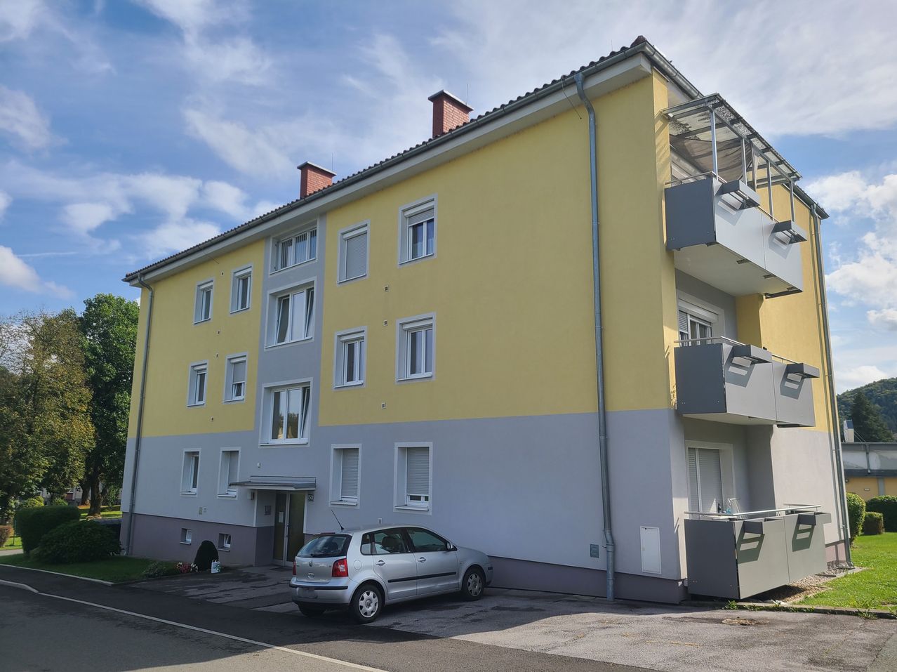 Ein gelbes und graues Mehrfamilienhaus mit mehreren Fenstern und Balkonen, ein silbernes Auto davor geparkt.
