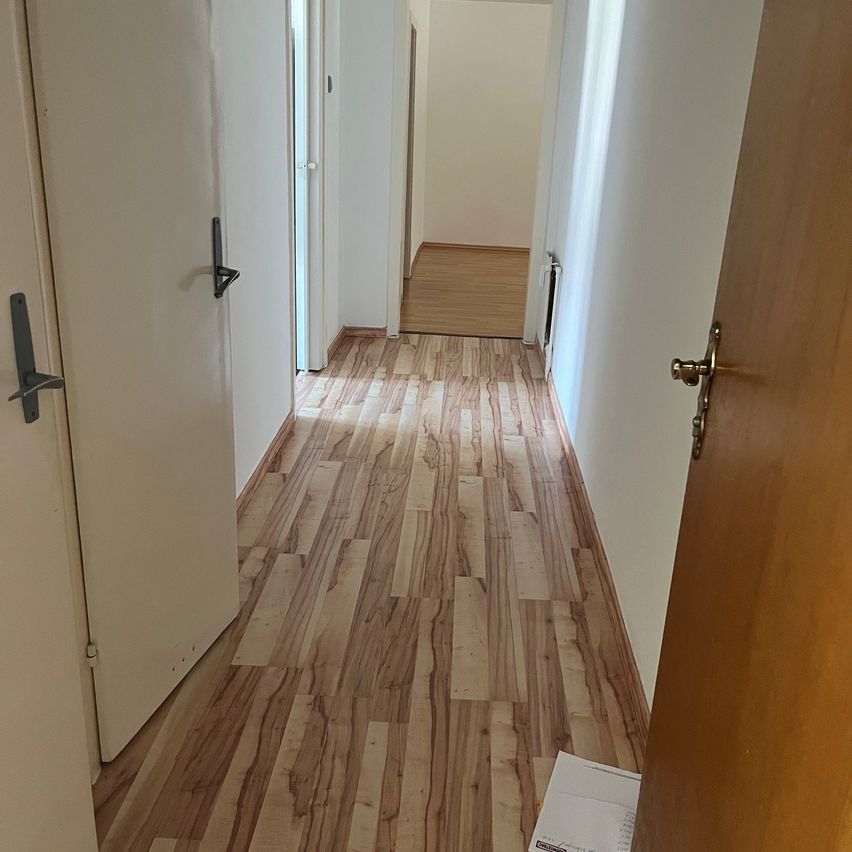 Ein leerer Flur mit Holzböden und weißen Wänden. Es gibt zwei geschlossene Türen auf der linken Seite und eine teilweise offene Tür auf der rechten Seite. Ein weißer Karton liegt auf dem Boden in der Nähe der offenen Tür.