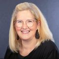 Ein Porträt einer Frau mit blonden Haaren, die eine Brille, Ohrringe und ein schwarzes Shirt trägt, lächelt in die Kamera.