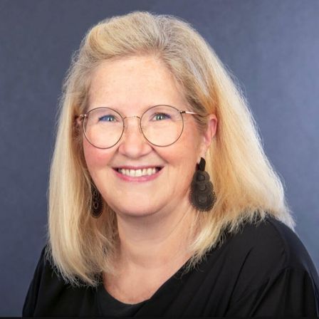 Ein Porträt einer Frau mit blonden Haaren, die eine Brille, Ohrringe und ein schwarzes Shirt trägt, lächelt in die Kamera.