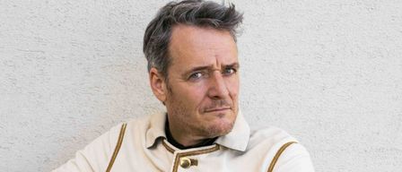 Ein Mann sitzt auf einem Holzstuhl und berührt seine Brust mit der linken Hand. Er trägt einen weißen Mantel mit Goldbesatz und ein schwarzes Innenhemd.