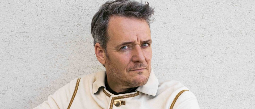 Ein Mann sitzt auf einem Holzstuhl und berührt seine Brust mit der linken Hand. Er trägt einen weißen Mantel mit Goldbesatz und ein schwarzes Innenhemd.