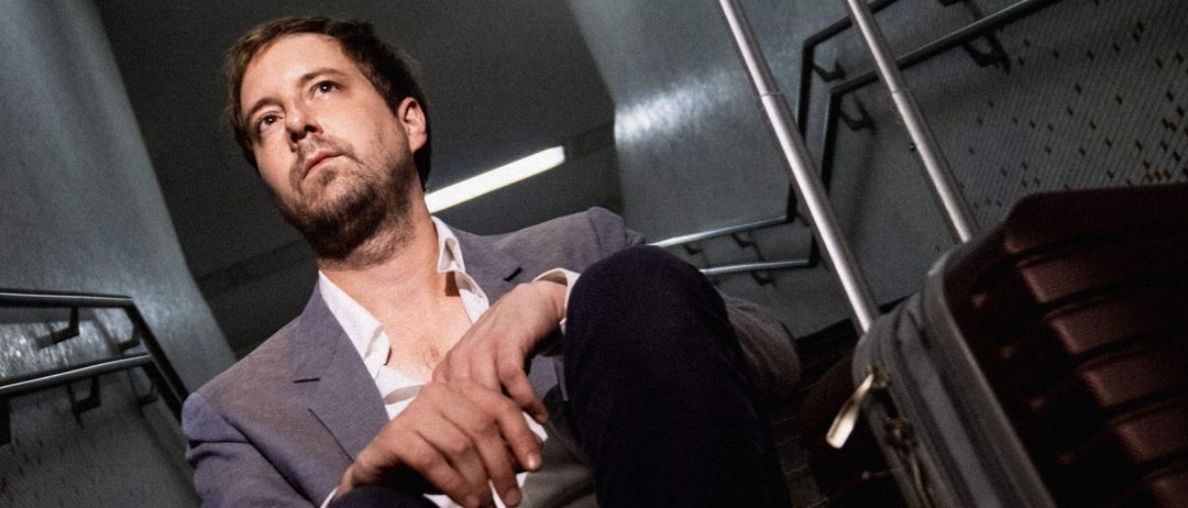 Ein Mann sitzt auf einer Treppe mit einem Koffer neben ihm. Er trägt einen Anzug und hat einen Bart. Die Treppe hat eine geflieste Wand und Metallgeländer.