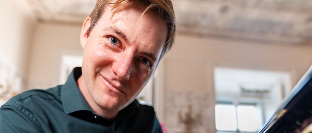 Ein Mann in einem schwarzen Hemd lächelt in die Kamera, vor einem Fenster und einem Spiegel stehend.