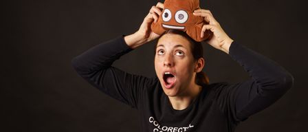 Eine Frau hält eine braune Plüsch-Kacke auf dem Kopf. Sie trägt ein schwarzes T-Shirt mit der Aufschrift 'Dickdarmkrebs würde nicht empfehlen'.