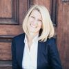 Eine Frau mit kurzen blonden Haaren lächelt in die Kamera. Sie trägt eine marineblaue Jacke über einem weißen Hemd und steht vor einer Holztür.