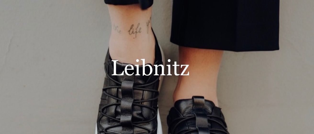 Nahaufnahme von schwarzen Schnürsenkel-Sneakers mit der Marke 'Leibnitz' und einer Tätowierung am Knöchel.