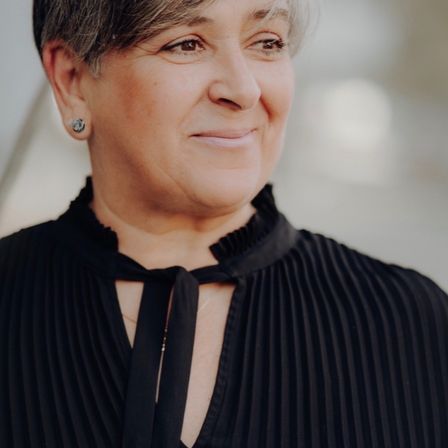 Eine Frau mit kurzen Haaren und einem schwarzen Kleid lächelt in die Kamera. Sie trägt einen silbernen Ohrring. Der Hintergrund ist unscharf.