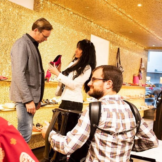 Ein Mann in einem grauen Anzug und ein anderer Mann in einem karierten Hemd schauen eine Frau an, die einen rosafarbenen High Heel hält. Sie sind in einem Schuhgeschäft mit golden glitzernden Wänden.