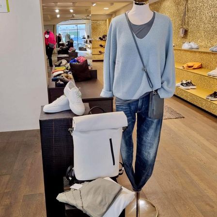 Ein Bekleidungsgeschäft zeigt eine Puppe mit einem hellblauen Strickoberteil, Jeans und einer weißen Tasche. Ein Tisch in der Nähe hat weiße Turnschuhe und eine weiße Rucksack.