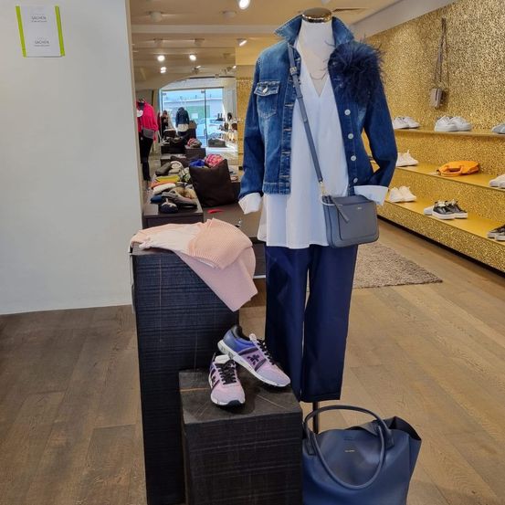 Ein Modegeschäft zeigt eine Puppe in einer Jeansjacke, einem weißen Oberteil, blauen Hosen und Sneakers. Eine blaue Handtasche und Schuhe befinden sich in den Regalen. Ein Schild an der Wand steht 'Sachsen'.