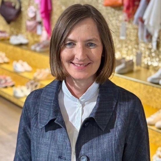 Eine Frau mit kurzen Haaren lächelt und steht vor einer Ausstellung von Schuhen und Taschen.