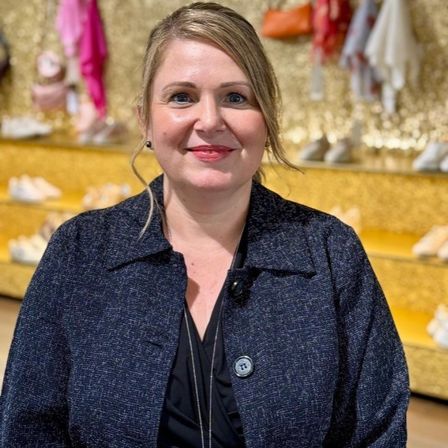Eine Frau mit blonden Haaren, die einen blauen Blazer trägt, steht vor einem Geschäft mit goldenen Regalen und verschiedenen ausgestellten Artikeln.
