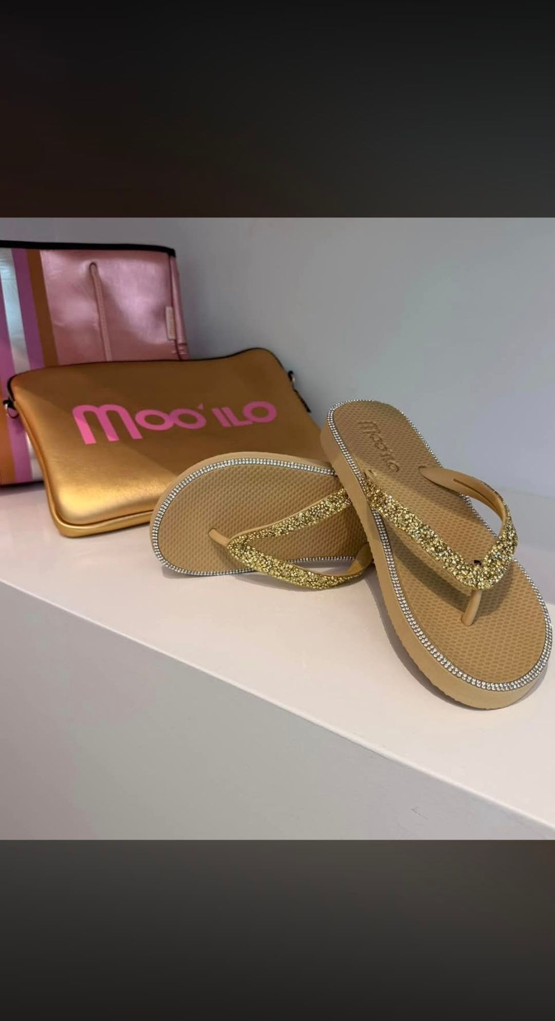 Zwei goldfarbene Sandalen mit Glitzerakzenten werden auf einer weißen Oberfläche ausgestellt. Dahinter befindet sich eine goldfarbene Tasche mit dem Wort 'Mooi'LO' in Pink geschrieben.