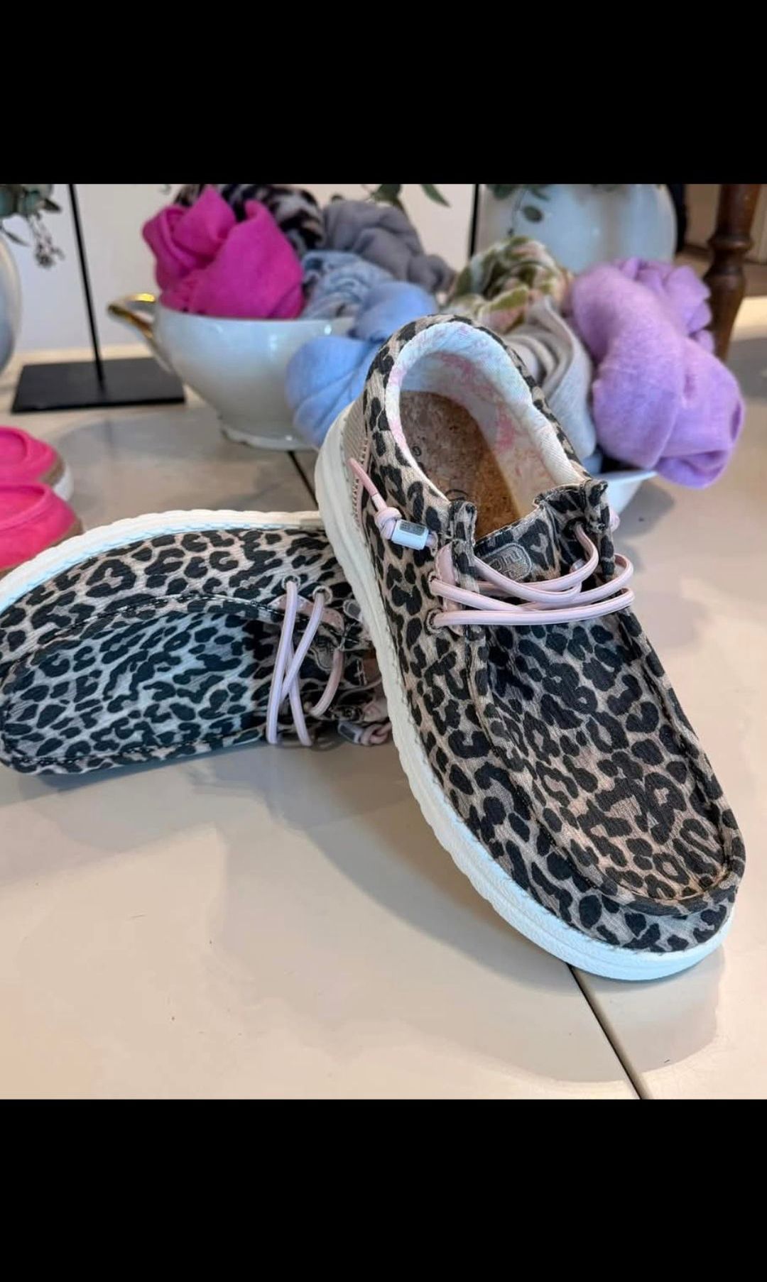 Ein Paar Leoparden-Print-Sneaker mit rosa Schnürsenkeln auf einer weißen Oberfläche, mit anderer Kleidung im Hintergrund.