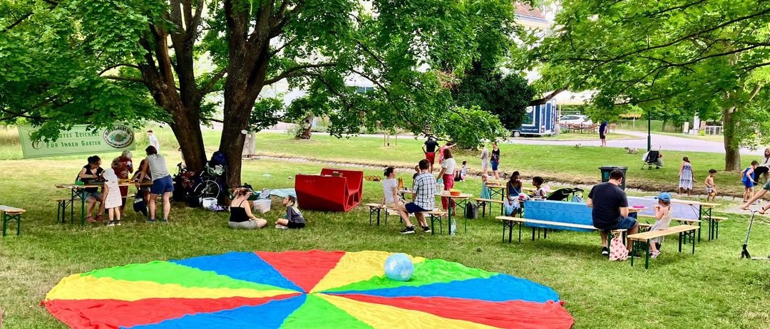 Eine Gruppe von Menschen in einem Park. Einige sitzen auf Bänken, andere stehen oder sitzen auf dem Rasen. Es gibt eine große bunte Decke auf dem Boden.
