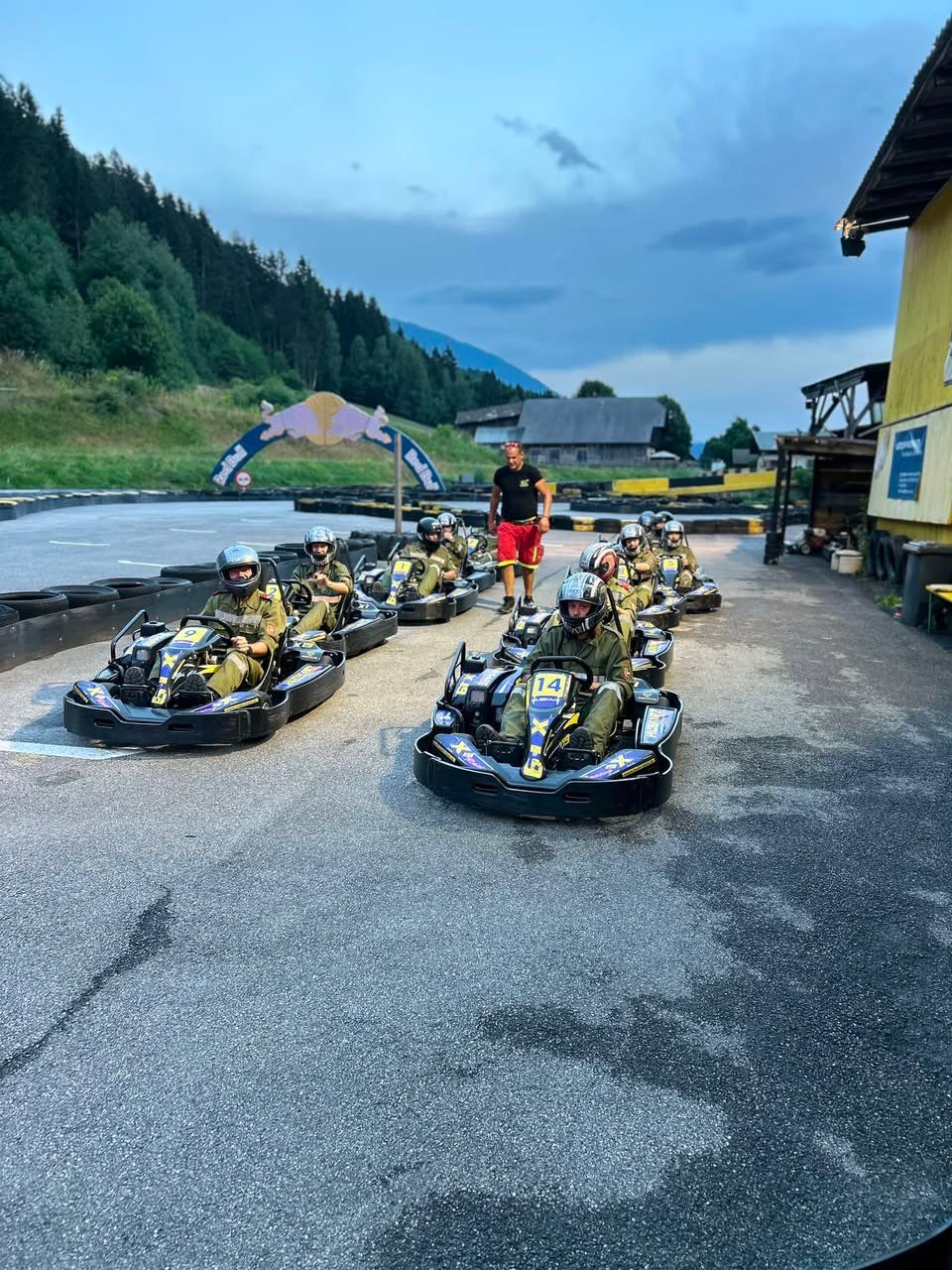 Eine Gruppe von Menschen in Helmen und Rennanzügen fährt Go-Karts auf einer Rennstrecke, ein Mann in einem schwarzen T-Shirt und roten Shorts steht in der Nähe.