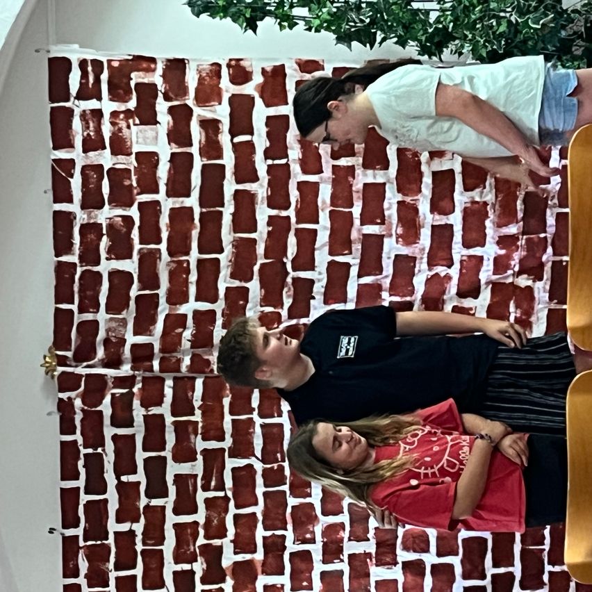 Drei Personen posieren vor einer roten Ziegelwand. Eine Person lehnt sich gegen die Wand, während die anderen beiden auf dem Boden liegen.