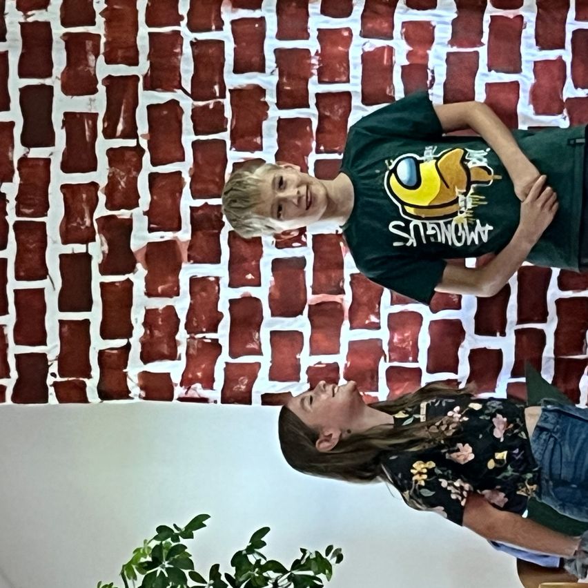 Zwei Personen, ein Junge in einem grünen T-Shirt und ein Mädchen in einem Blumenshirt, lächeln und stehen vor einer Backsteinmauer.