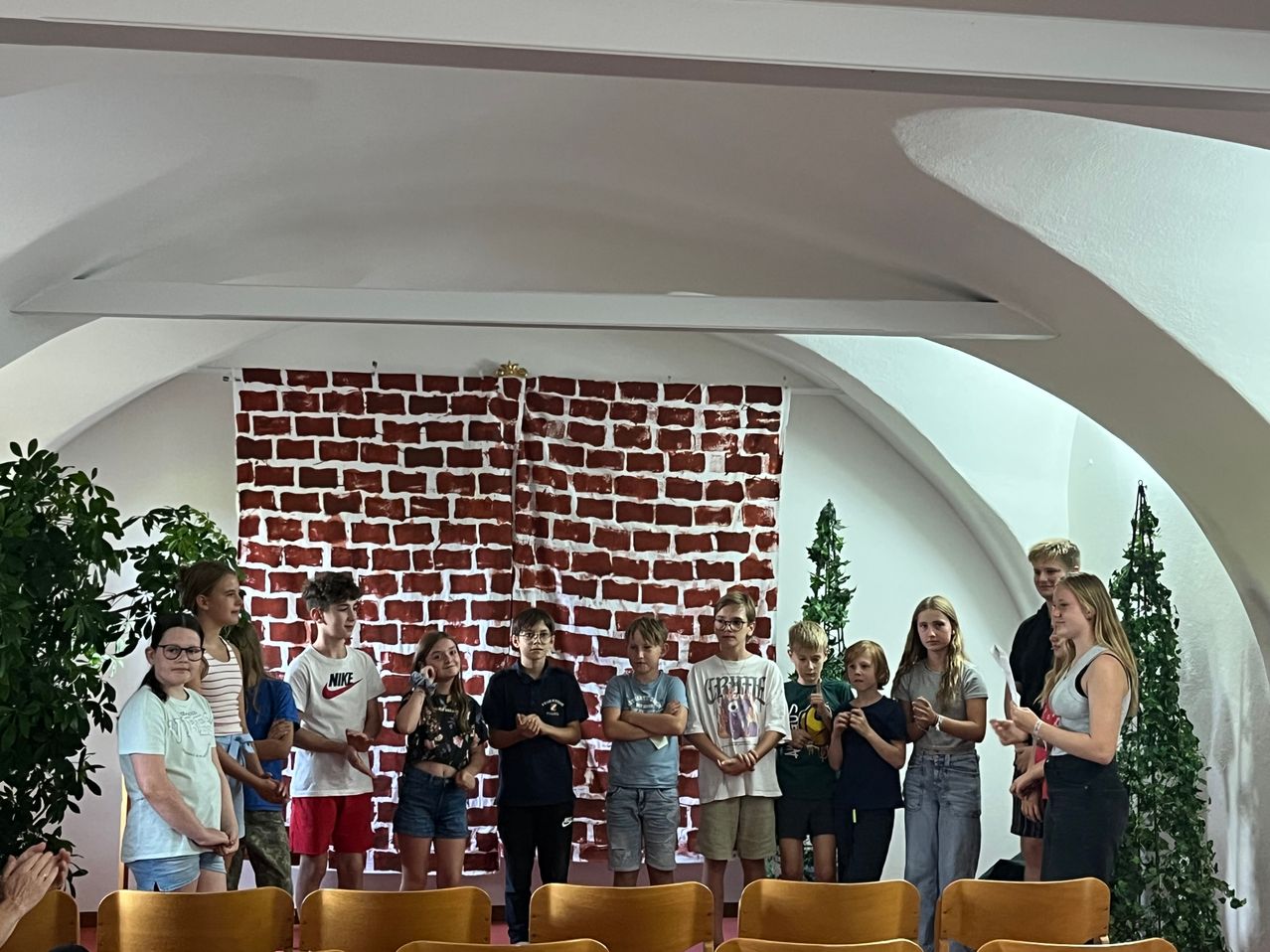 Eine Gruppe von Kindern und Jugendlichen steht vor einer roten Ziegelwand. Sie scheinen an einem Chor oder einer Gesangsaufführung teilzunehmen. Die Umgebung ist drinnen mit hölzernen Stühlen in Reihen angeordnet.
