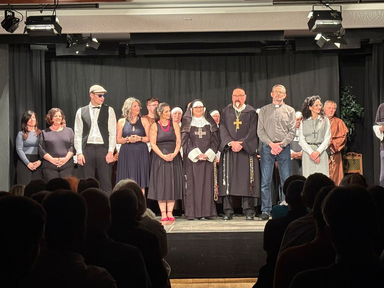 Eine Gruppe von Darstellern steht auf der Bühne vor einem Publikum. Zwei Männer tragen eine Brille und eine Fliege, während zwei Frauen rote Halsketten tragen. Eine Nonne und ein Priester stehen in der Mitte.