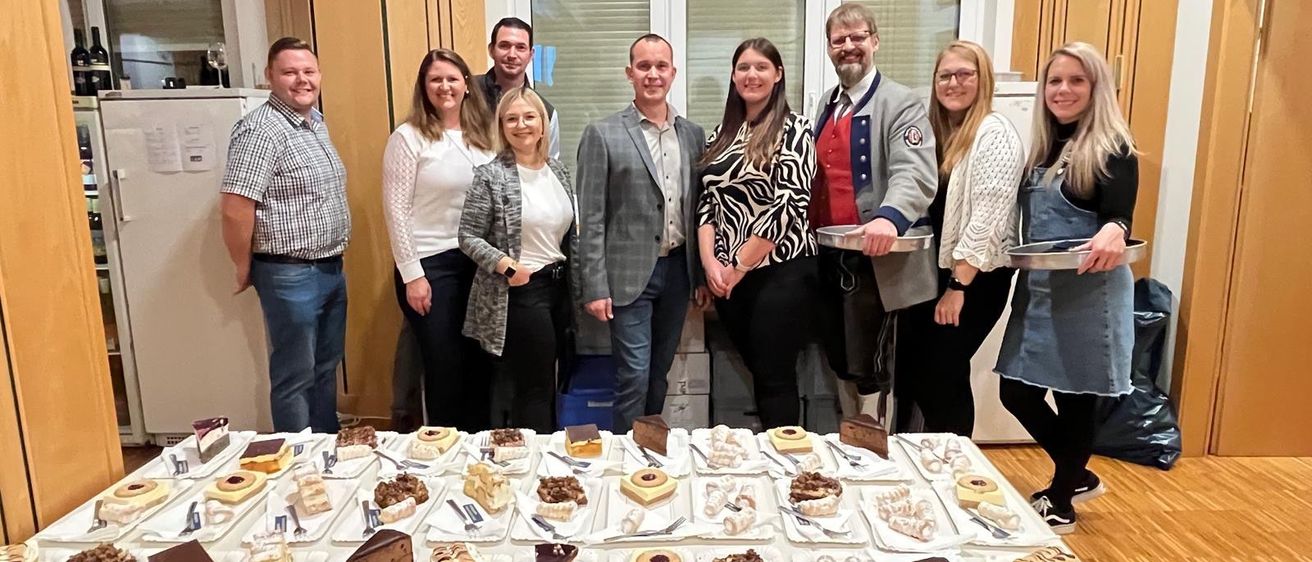 Eine Gruppe von Menschen steht hinter einem Tisch voller verschiedener Desserts und posiert für ein Foto. Sie tragen Freizeitkleidung. Hinter ihnen befinden sich Fenster und eine Holzwand.