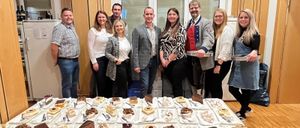 Eine Gruppe von Menschen steht hinter einem Tisch voller verschiedener Desserts und posiert für ein Foto. Sie tragen Freizeitkleidung. Hinter ihnen befinden sich Fenster und eine Holzwand.