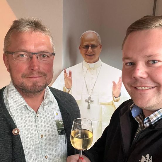 Zwei Männer lächeln für ein Foto mit einem großen Plakat von Papst Franziskus im Hintergrund. Ein Mann hält ein Glas Weißwein.